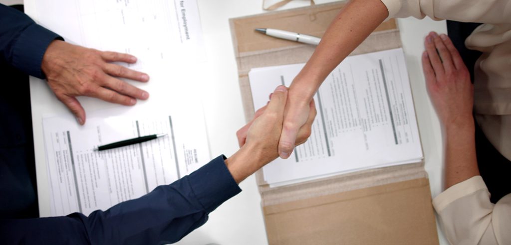 Two people shaking hands over paperwork at a table in an office.